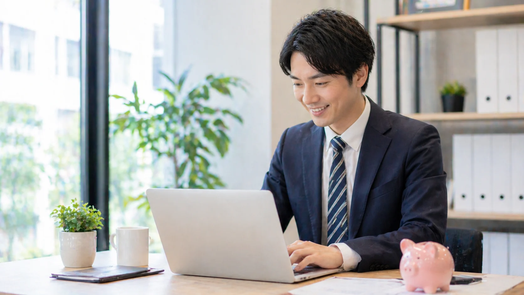 明るいオフィスでスーツ姿の男性が貯金箱の置かれたデスクで笑顔でノートパソコンを操作している。